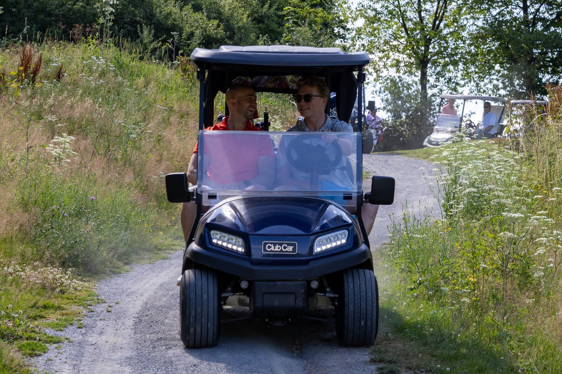 Golfcart mit zwei Insassen auf einem Weg zwischen hohen Wildblumenwiesen
