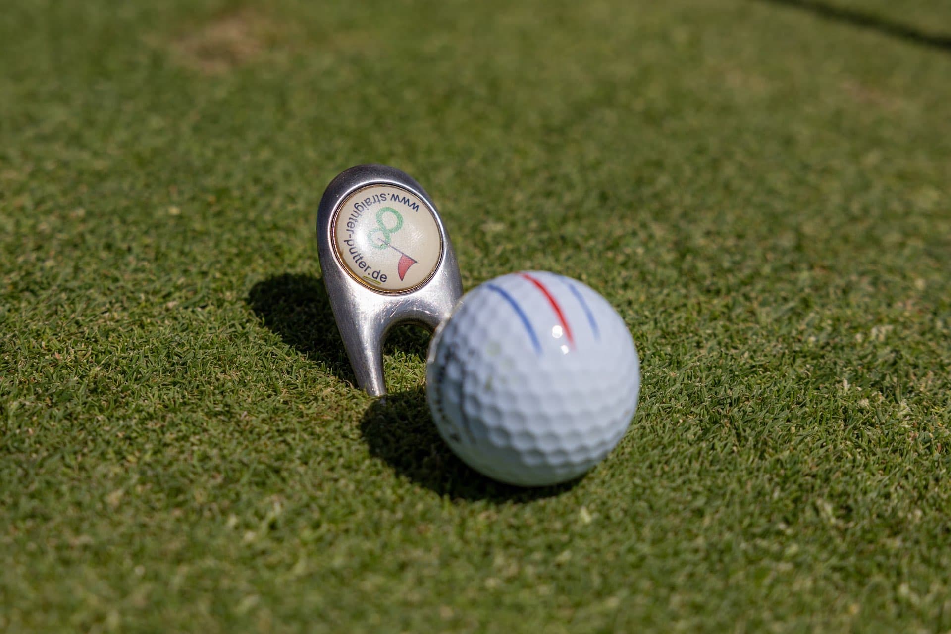 Pitchgabel mit rundem Logo-Einsatz steckt neben einem Golfball im Grün