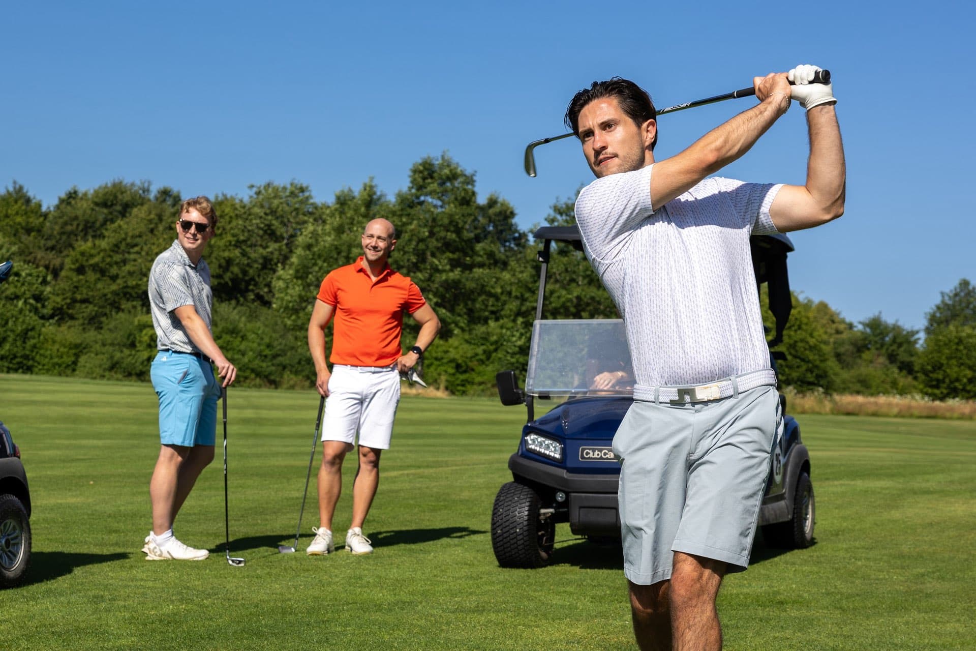 Ein Spieler im Durchschwung, zwei Mitspieler schauen am Golfcart zu