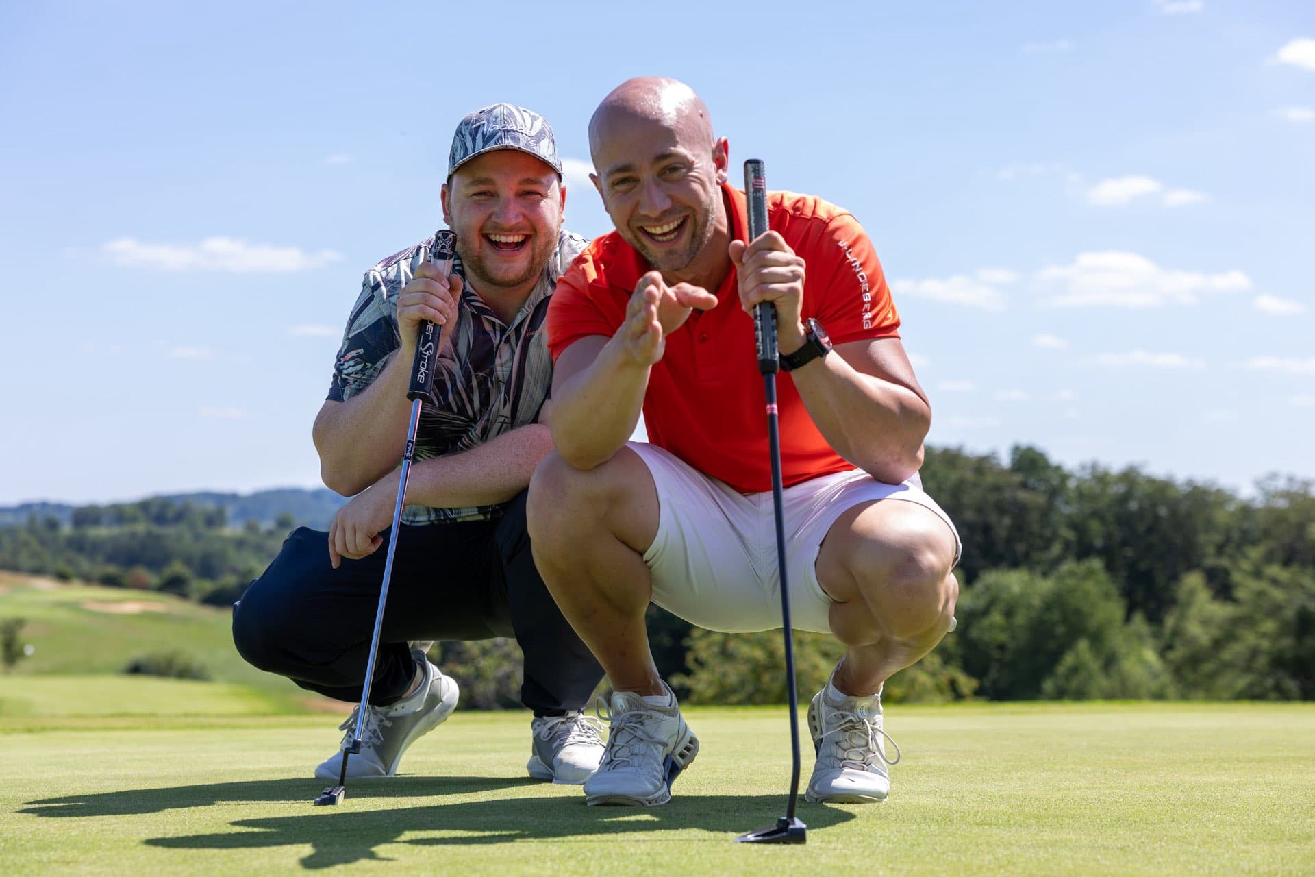 Zwei Golfer hocken lachend auf dem Grün, aufgestützt auf ihre Putter