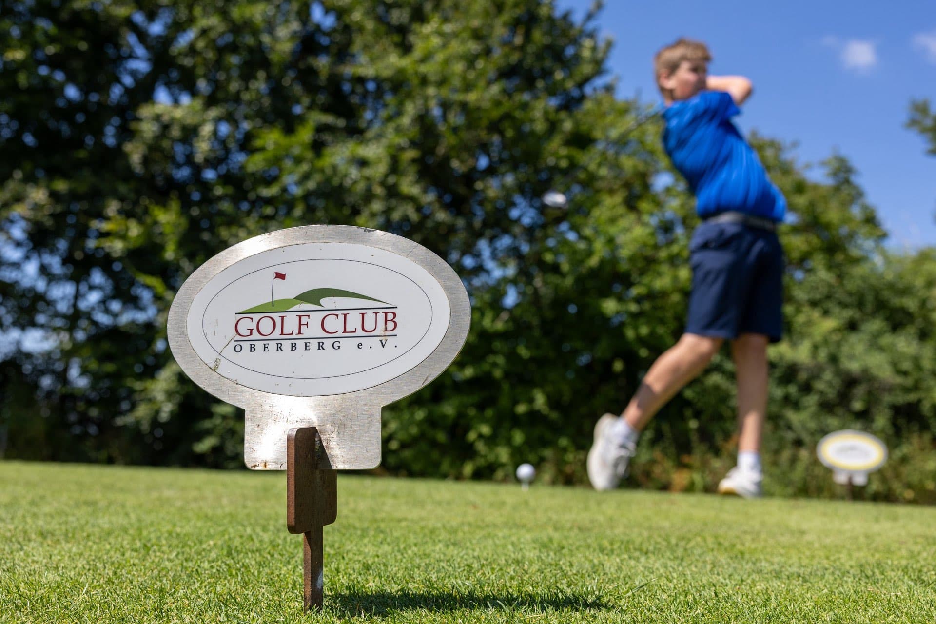 Abschlagschild mit dem Logo des Golf Club Oberberg, dahinter ein Jugendlicher