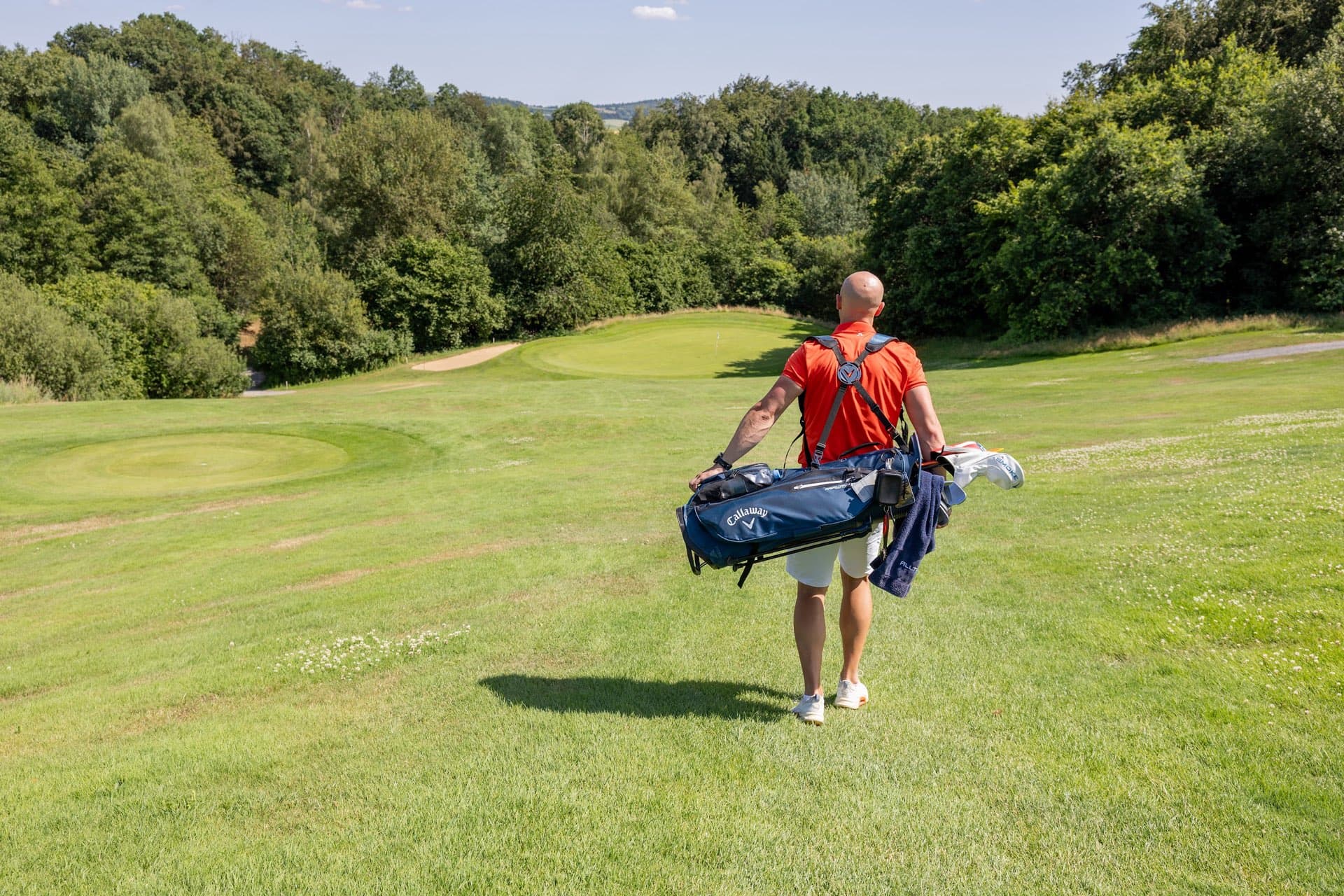 Golfer mit Bag über der Schulter geht einen Hang hinunter zum Grün
