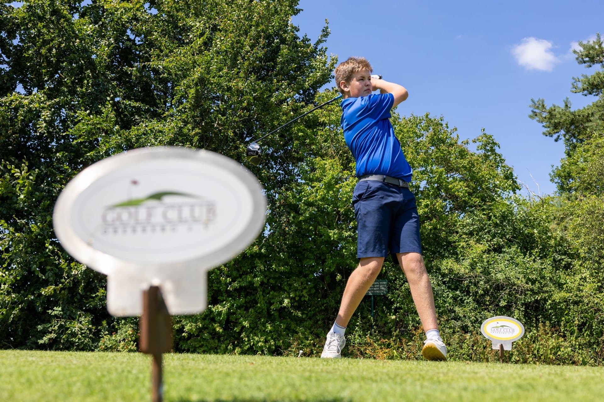Abschlagschild des Golf Club Oberberg, dahinter schwingt ein Jugendlicher aus